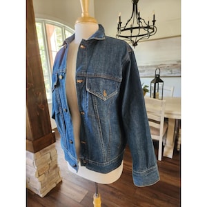 May include: A classic blue denim jacket with a button-down front, two chest pockets, and gold-colored buttons. The jacket is displayed on a mannequin, showcasing its traditional design and fit. The jacket is a medium wash denim.