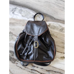 May include: A dark brown leather backpack featuring a rounded top flap with a brass-toned buckle closure, a drawstring opening, a top carry handle, and thin shoulder straps. The bag is accented with thin tan piping along the edges.