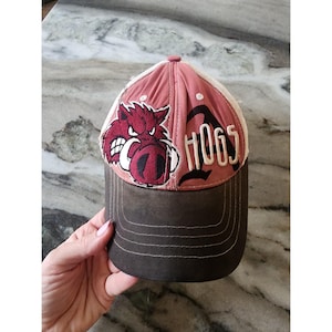 Könnte beinhalten: Eine braune und rosa Baseballkappe mit Mesh-Rückseite. Die Kappe zeigt eine rot-schwarze Grafik eines Wildschweins und die Zahl "1065" in Weiß. Der Schirm ist braun und die Krone rosa.