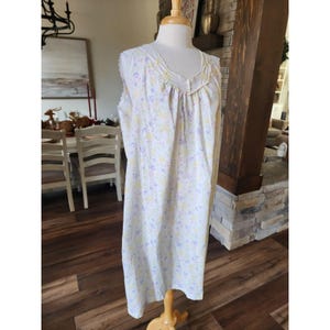 May include: A white nightgown with a floral pattern in yellow and purple is displayed on a mannequin. The nightgown has a scoop neckline with lace trim and a loose, flowing fit. The fabric appears to be lightweight and comfortable, suitable for sleepwear.