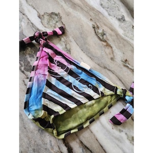 May include: A vibrant bikini bottom with tie sides. The fabric displays a striped pattern in black, blue, pink, and white, with a green interior lining. The bikini bottom has a decorative cursive letter design.