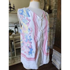 Puede incluir: Top blanco sin mangas con estampado floral en rosa, azul y amarillo. El top tiene cuello en V, botones decorativos y un diseño a rayas rosas en la parte delantera. El dobladillo inferior está adornado con encaje blanco.
