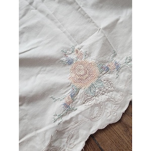 May include: White fabric with a floral embroidery design. The embroidery features a central pink rose surrounded by blue and green accents. The edge of the fabric has a scalloped design with embroidered details.