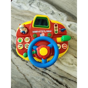 May include: A VTech See Me Go Driver toy featuring a blue steering wheel, a red dashboard with a small mirror, a green gear shift, and various colourful buttons with icons including a gas pump, a stop sign, and a small dog figure on the left side.