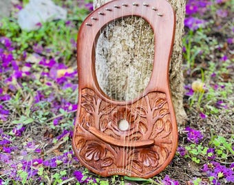 May include: A handcrafted wooden lyre with intricate carvings of leaves and branches. The instrument has multiple strings and is resting against a tree trunk, surrounded by purple flowers and green grass.