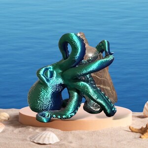 May include: A teal blue octopus figurine with a textured surface, holding a clear glass bottle. The octopus is sitting on a light brown circular base.