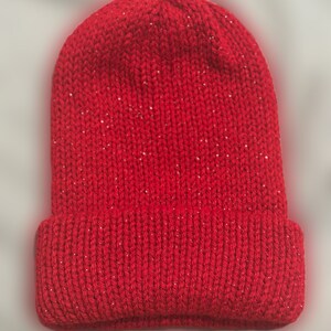 May include: A bright red knitted beanie hat with a turned-up brim. The hat is made from a textured yarn with tiny silver sparkles woven in. This warm winter hat is ideal for cold weather, and measures approximately 20 cm in height.