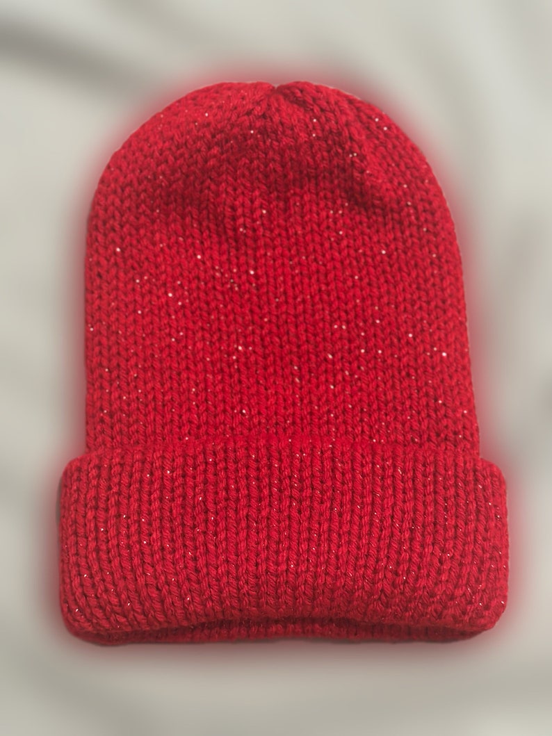 May include: A bright red knitted beanie hat with a turned-up brim. The hat is made from a textured yarn with tiny silver sparkles woven in. This warm winter hat is ideal for cold weather, and measures approximately 20 cm in height.