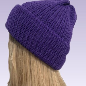 May include: A knitted beanie hat in a deep purple colour. The hat is made from a thick, textured yarn and features a folded brim. The hat is shown on a person with blonde hair. This winter hat is a stylish accessory.