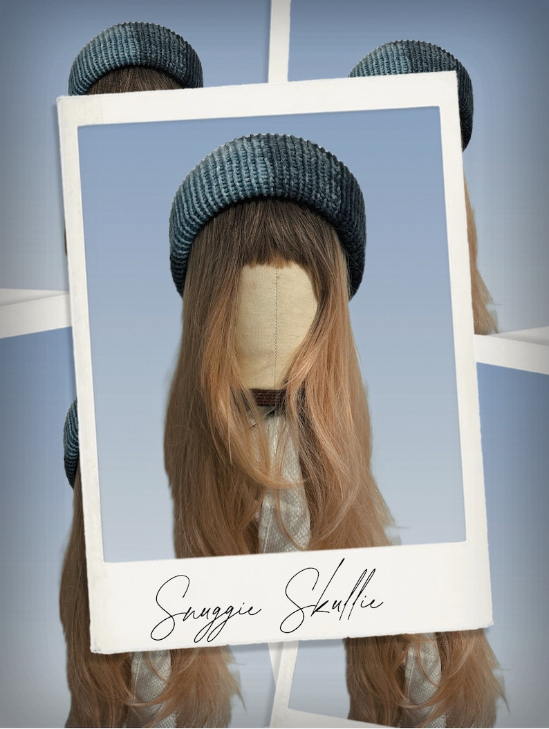 May include: A blue and grey knitted beanie hat on a mannequin head with long, light brown hair. The image is framed within a white border, with the words "Snuggie Skullie" in cursive script at the bottom.
