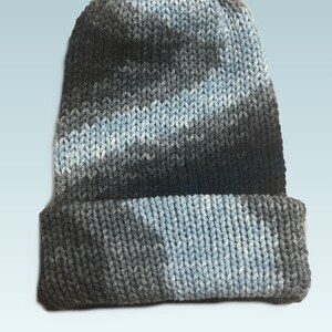 May include: A knitted beanie hat in shades of grey and light blue. The hat has a folded brim and a textured knit pattern. The colour transitions create a subtle gradient effect, adding visual interest to the winter accessory.