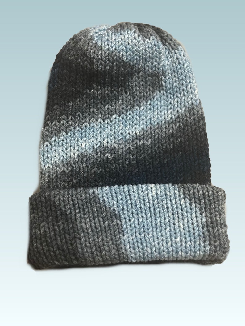 May include: A knitted beanie hat in shades of grey and light blue. The hat has a folded brim and a textured knit pattern. The colour transitions create a subtle gradient effect, adding visual interest to the winter accessory.