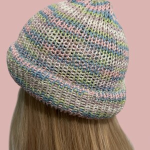 May include: A knitted beanie hat in pastel shades. The hat has a mesh-like pattern, incorporating pink, blue, green, and white threads. The brim is turned up, and the hat is viewed from the side.