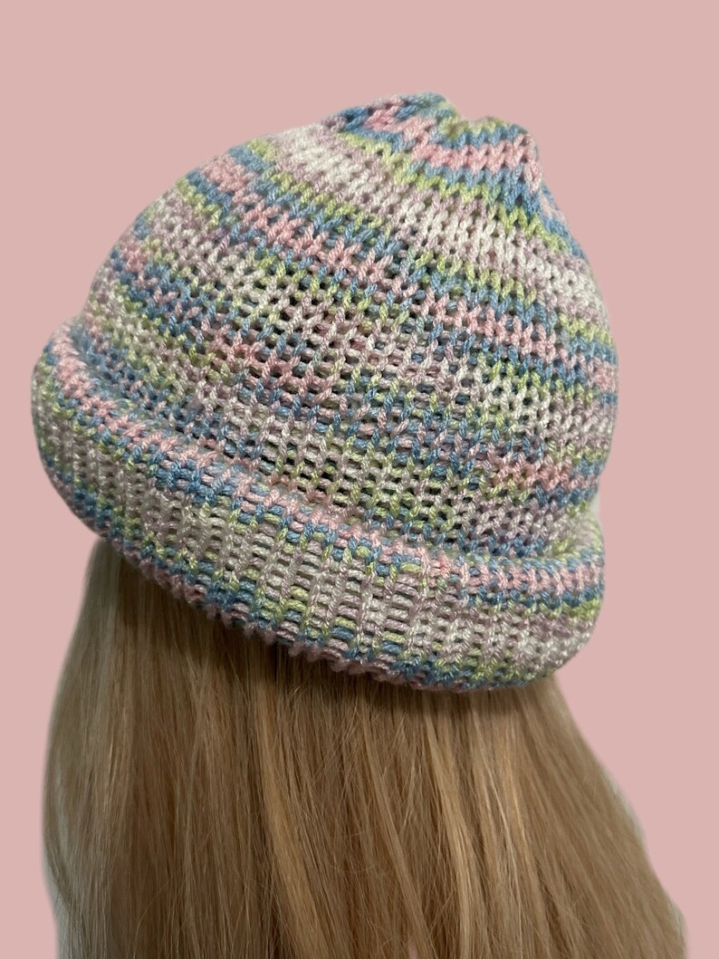 May include: A knitted beanie hat in pastel shades. The hat has a mesh-like pattern, incorporating pink, blue, green, and white threads. The brim is turned up, and the hat is viewed from the side.