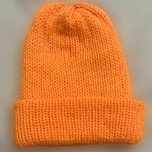 May include: A vibrant orange knitted beanie hat. The hat has a folded-over brim and is made from a thick, textured yarn. This solid-coloured beanie is suitable for cooler temperatures.