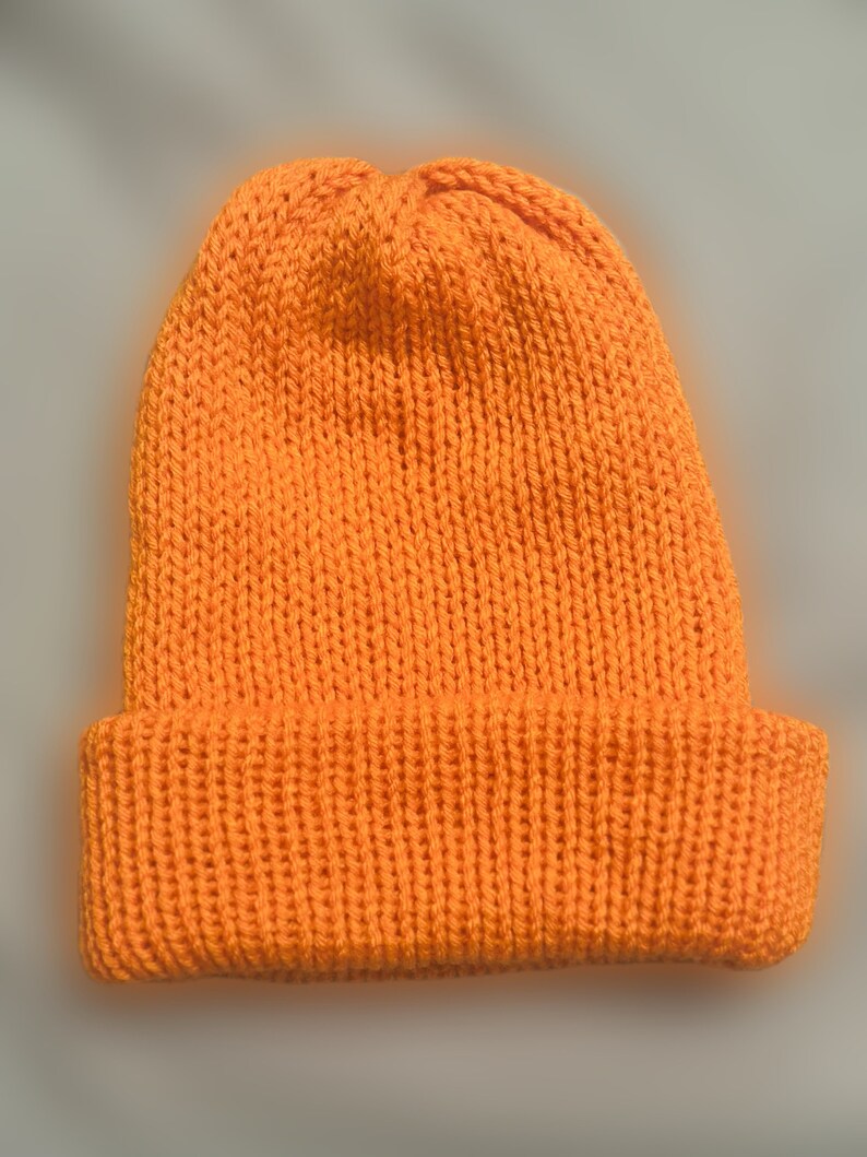 May include: A vibrant orange knitted beanie hat. The hat has a folded-over brim and is made from a thick, textured yarn. This solid-coloured beanie is suitable for cooler temperatures.