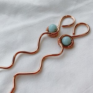 May include: A pair of copper hair pins with a wavy design. Each pin features a light blue gemstone wrapped in copper wire.