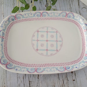 Könnte beinhalten: Rechteckige weiße Keramik-Servierplatte mit dekorativem Rand in Rosa, Blau und Lila. Die Mitte zeigt ein kreisförmiges Design mit einem Gittermuster und floralen Akzenten. Eine charmante Ergänzung für jede Tischdekoration.