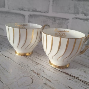 May include: Two white teacups with gold detailing. Each cup features gold vertical stripes and a gold base. The text "Congratulations 50" is printed in gold inside the cup, perfect for a celebration.