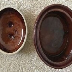 Puede incluir: Cazuela de cerámica marrón de dos piezas, de forma ovalada. La tapa es de un tono marrón más oscuro que la base, que tiene un borde más claro. La tapa tiene dos pequeños agujeros. Para hornear o servir.