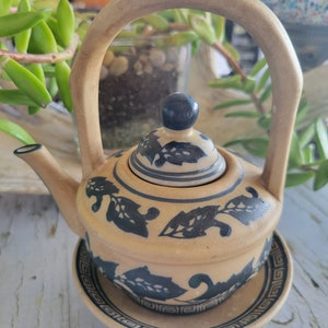 Puede incluir: Una tetera de cerámica beige con un platillo a juego. La tetera presenta un asa curva y una tapa con un pomo negro. Patrones de hojas azul oscuro decoran el cuerpo y la tapa. El platillo tiene un borde azul a juego.
