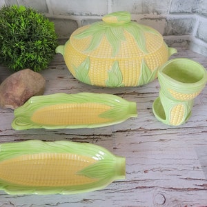 Puede incluir: Un conjunto de utensilios de cocina de cerámica con temática de maíz. Incluye una fuente para servir con tapa, dos bandejas y un soporte para utensilios en forma de mazorca de maíz. Las piezas son amarillas y verdes, con un diseño detallado de mazorca.