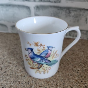 May include: White ceramic mug with a gold rim and handle. The mug features a detailed illustration of two blue birds perched on a branch with autumn-colored leaves. A classic design for a coffee or tea cup.