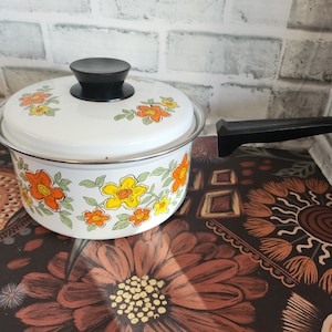 Peut inclure: Une casserole en émail blanc avec un couvercle, ornée de motifs floraux orange et jaunes. Elle possède une poignée noire et un bouton noir sur le couvercle. Article de cuisine vintage.