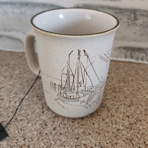 Puede incluir: Taza de cerámica beige moteada con borde y asa marrones. La taza presenta un dibujo lineal negro detallado de un velero atracado en un muelle. Adecuado para bebidas frías o calientes.