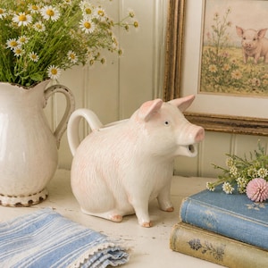 May include: A ceramic pig-shaped pitcher with a glossy white finish and soft pink accents on the ears, snout, and back. The pitcher features a curved handle on the back and an open mouth spout, designed in a realistic animal form with textured details.