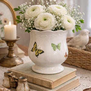 Könnte beinhalten: Eine weiße Keramikvase mit Schmetterlingsillustrationen enthält weiße Blumen und Grünzeug. Die Vase steht auf alten Büchern, mit einer Kerze und dekorativen Gegenständen im Hintergrund. Die Vase ist etwa 18 cm hoch.