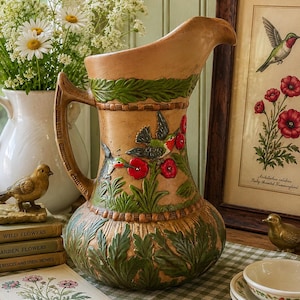 May include: A tan ceramic pitcher featuring a raised relief design of green leaves and hummingbirds feeding on red flowers. The pitcher has a curved handle and a wide spout, with decorative bands encircling the top and bottom sections.