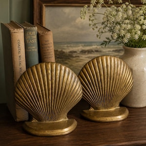 May include: A pair of gold-toned metal bookends shaped like large, fan-like seashells with prominent vertical ridges. Each bookend features a curved, semi-circular base that provides stability for holding books upright on a flat surface.
