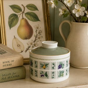 May include: A white ceramic sugar bowl with a lid, featuring a repeating grid pattern of various fruits and leaves around the exterior. The lid is a solid sage green color with a white knob. The bowl has a thin gold rim and a green leaf border.