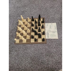 May include: A chess set with black and cream-coloured pieces arranged on a brown and tan chequered board. A printed paper with the word "Renaissance" is next to the board. The chess pieces are in various shapes.