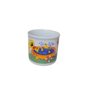 Puede incluir: Taza de cerámica blanca con una colorida ilustración de patos amarillos jugando en una bañera. El diseño incluye una escena de agua azul, un salvavidas rojo y blanco y hierba verde.