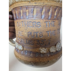May include: A handmade ceramic mug with a brown and tan speckled glaze. The mug features carved text that reads "Gathers the waters, He puts the deeps, Psalms 33:7." The mug has a handle and decorative floral accents.