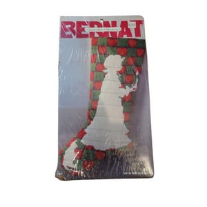 May include: A vintage Bernat Quic stitch stocking craft kit featuring a girl holding a candle in front of a red and green heart pattern. The kit includes instructions and yarn.