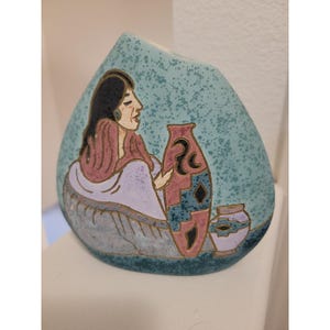 Könnte beinhalten: Eine türkisfarbene Keramikvase mit einer bemalten indianischen Frau und zwei dekorativen Vasen. Die Frau trägt einen rosa Schal und hat schwarzes Haar. Die größere Vase ist rosa und blau, die kleinere lila und blau.