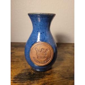 Könnte beinhalten: Kleine blaue Keramikvase mit rundem Körper und schmalem Hals. Ein kreisförmiges, braunes Emblem mit einem Kronenmuster und dem Text "CHARLOTTE NC" ist auf der Vorderseite angebracht. Die Vase steht auf einer Holzoberfläche.
