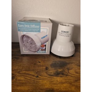Puede incluir: Un difusor de cabello blanco Conair Euro Style y su caja. La caja tiene las palabras "Euro Style Diffuser" y "Lifts hair with its fingers maintains curl without frizzing for all hair types". El difusor está diseñado para levantar el cabello y mantener los rizos.