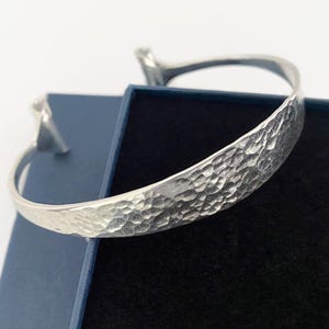 May include: A silver hammered metal cuff bracelet. The bracelet has a textured surface and a curved design. The bracelet is displayed on a dark blue box with a black interior. Jewellery.