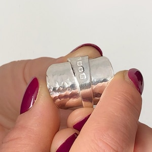 May include: A silver ring with a hammered finish and a rectangular band. The ring has a wide, curved design and is held between fingers. The nails are painted burgundy.