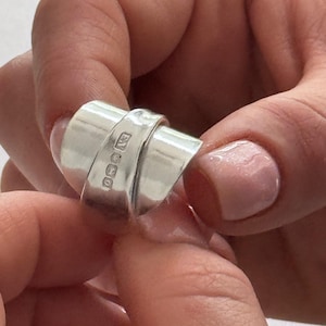May include: A wide, polished silver ring featuring a smooth, overlapping band design. The surface of the ring displays a series of small, rectangular hallmarks stamped into the metal, indicating its composition and origin in a clear, metallic finish.