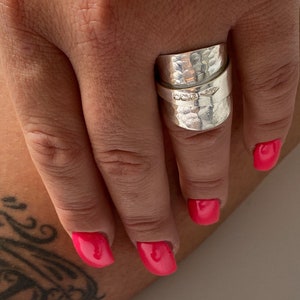May include: A silver ring with a hammered texture and a wrap-around design. The ring has a bright, reflective finish. The ring is worn on a finger with bright pink nail polish.