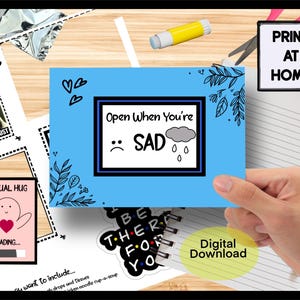 May include: A blue card with the text "Open When You're SAD" and a sad face and rain cloud illustration. The image also includes a "Virtual Hug" card, a "Print at Home" sign, and a "Digital Download" sticker.