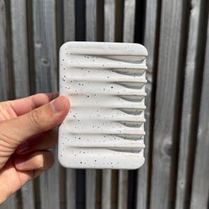 May include: A rectangular white soap dish featuring a speckled pattern of small black dots across its surface. The dish has a series of parallel horizontal ridges designed to elevate an object, with rounded corners and a smooth, matte finish.
