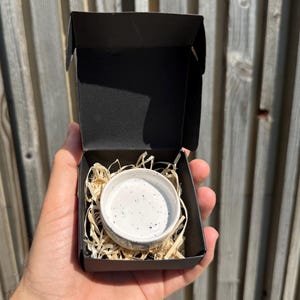 May include: A small, round, white ceramic bowl with black speckles nestled inside a black cardboard gift box. The bowl is surrounded by light-colored wood shavings used as protective packaging material within the open container.