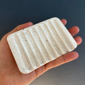 May include: A rectangular white ceramic soap dish featuring a speckled pattern with small black dots. The surface of the dish is designed with a series of parallel, raised ridges that run across the width, creating channels for drainage.
