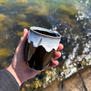 Könnte beinhalten: Eine schwarze Keramiktasse mit weißer, getropfter Glasur am Rand. Die Tasse wird in einer Hand gehalten, mit einem verschwommenen Hintergrund aus Wasser und Felsen.